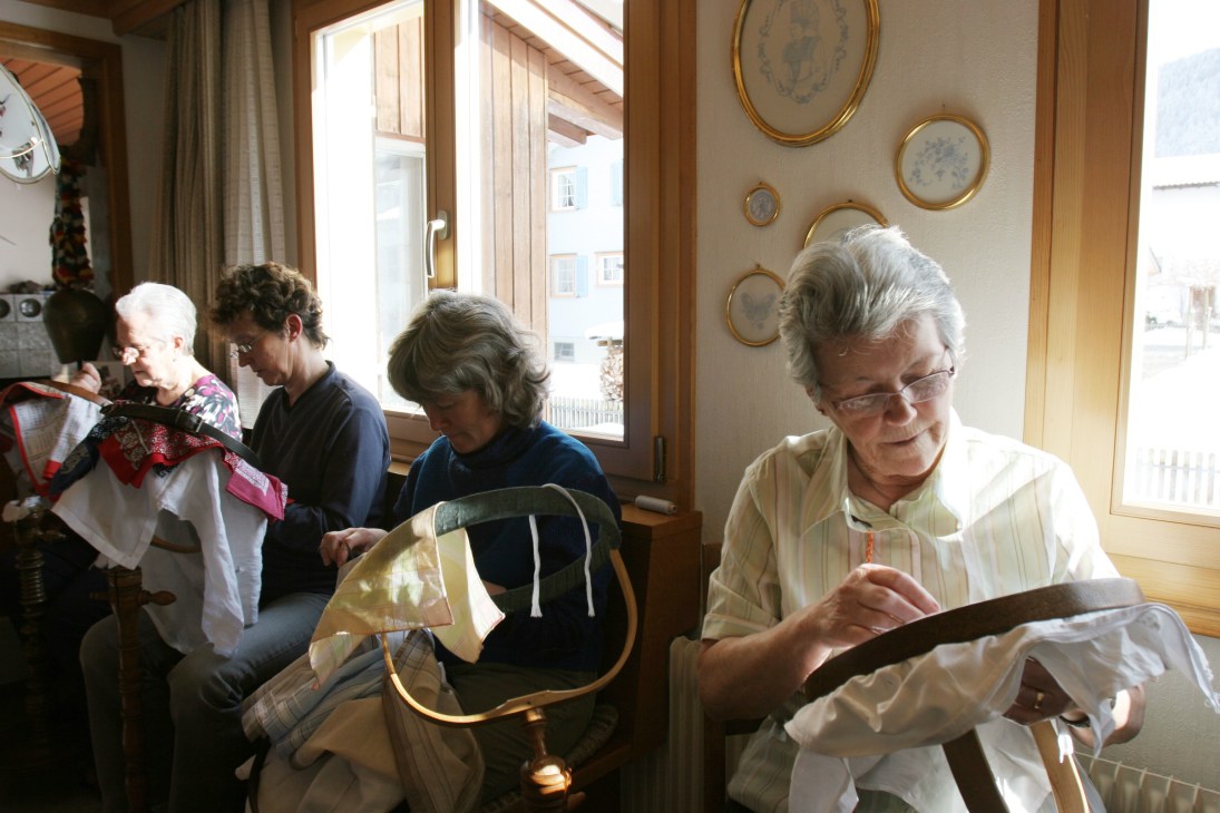 Elderly seamstresses working on embroidered linens in a bright workshop with framed needlework displayed on walls.