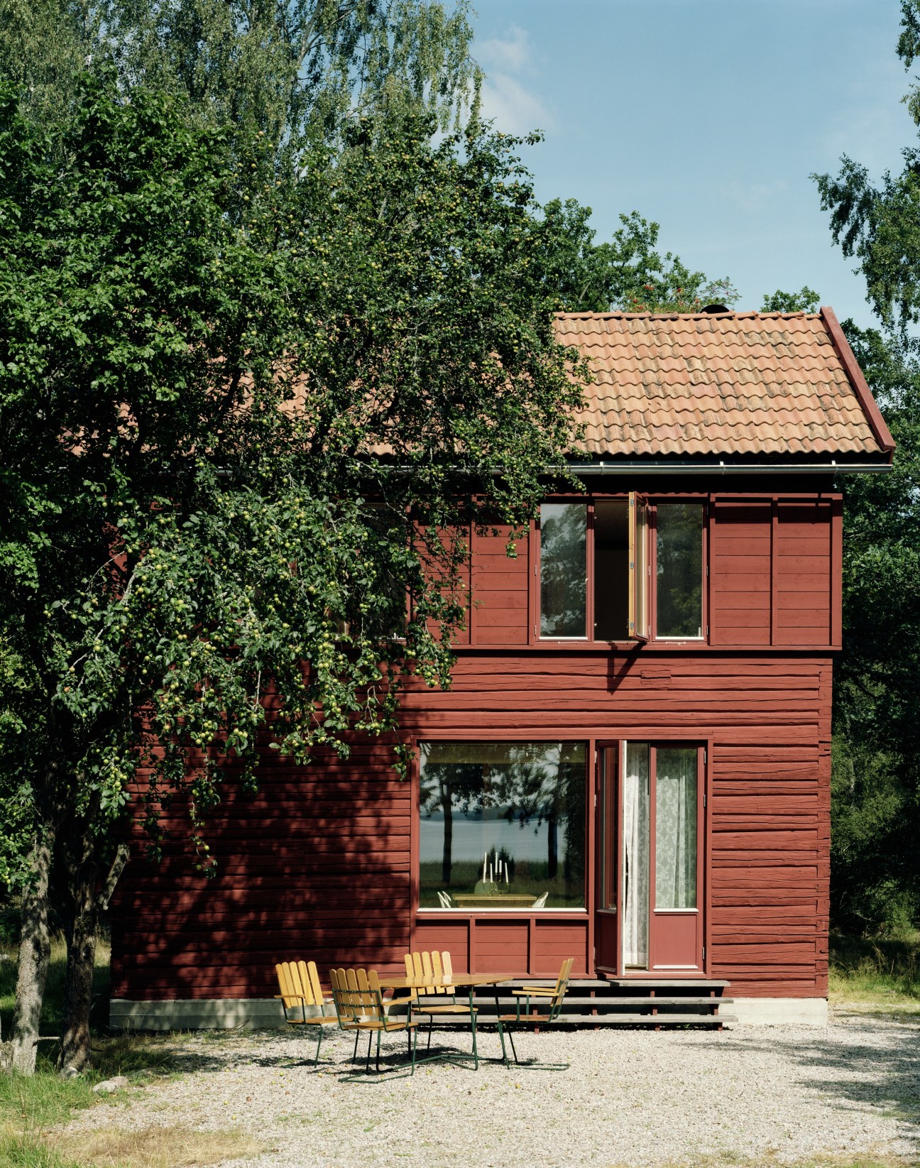 Red wooden summerhouse surrounded by birch trees with yellow outdoor chairs on a gravel patio