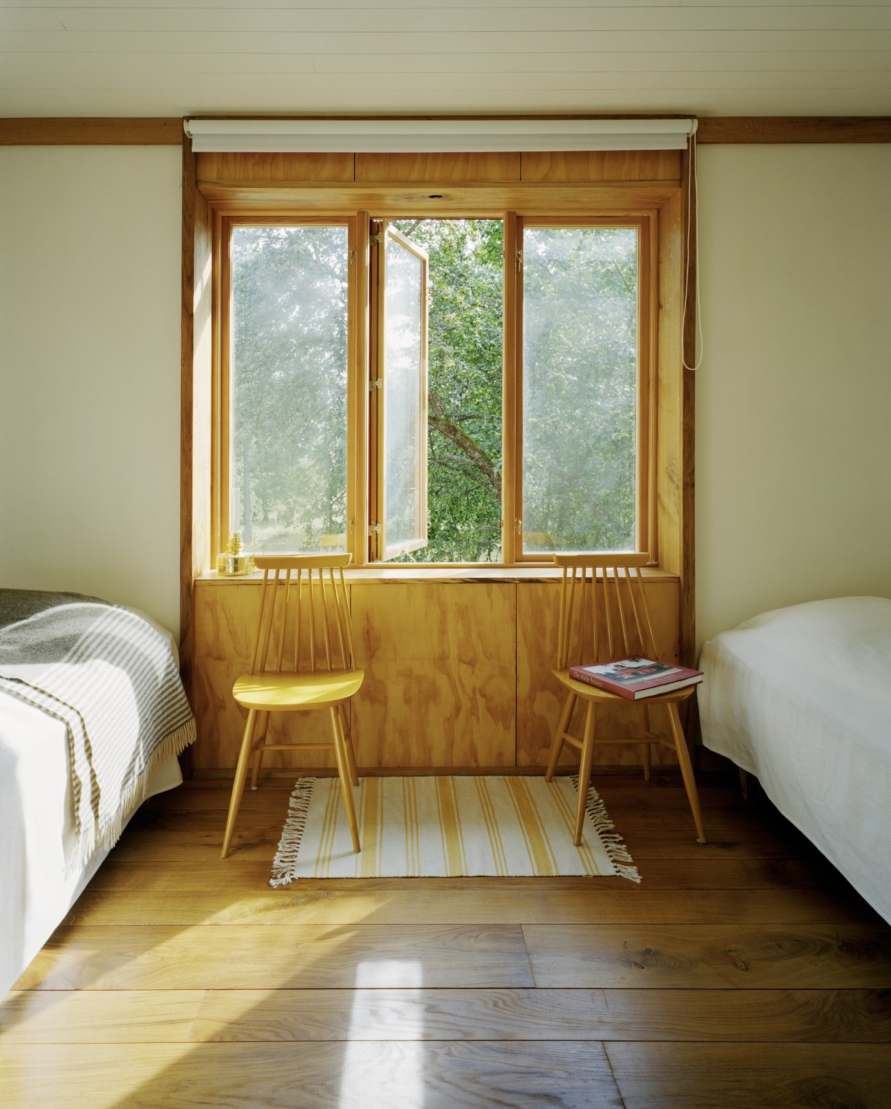 Children's bedroom with two beds flanking wooden-framed windows overlooking green woodland.