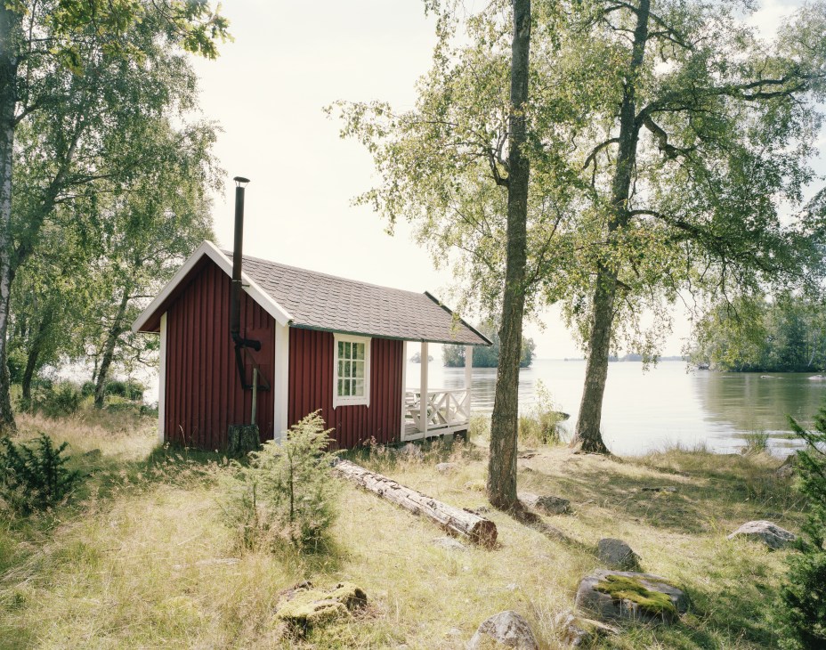 Red wooden sauna building with white porch situated on grassy ground between birch trees beside a calm lake.