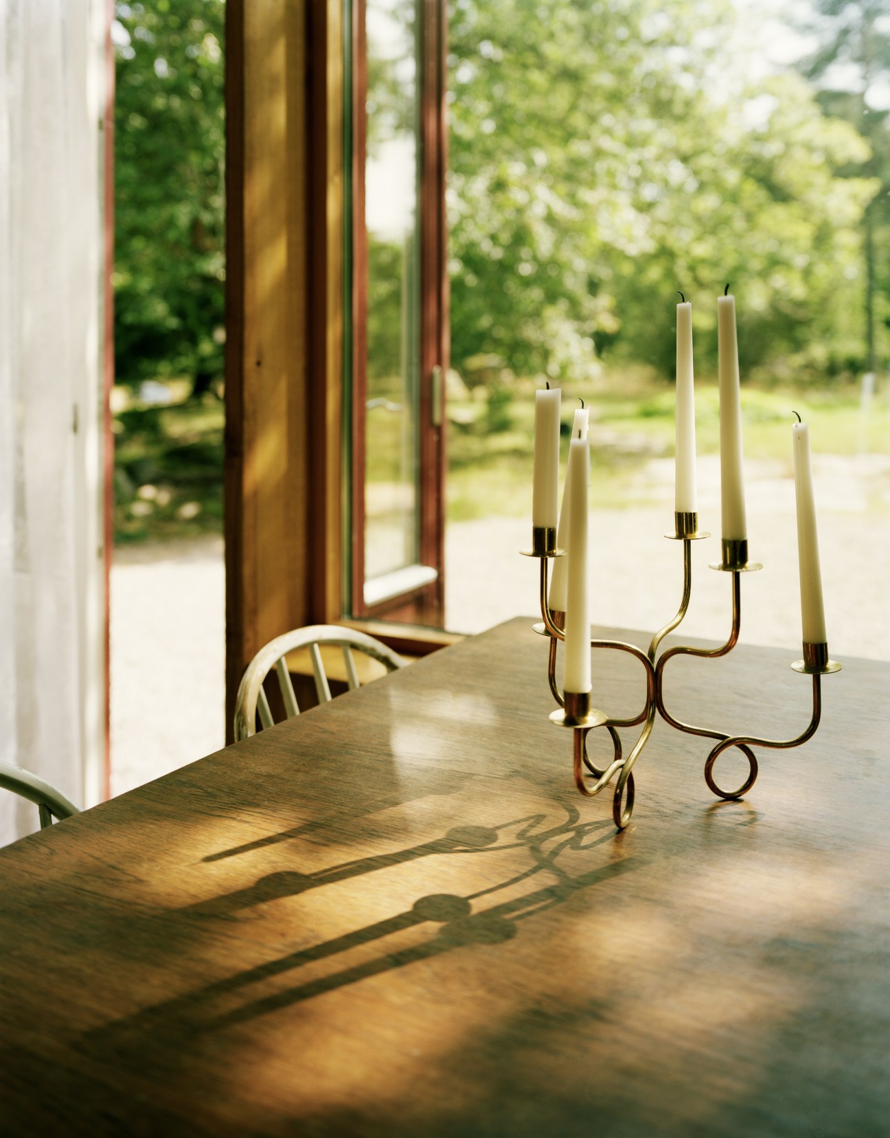 Brass candelabra with lit cream candles on a wooden dining table with large windows overlooking green landscape.