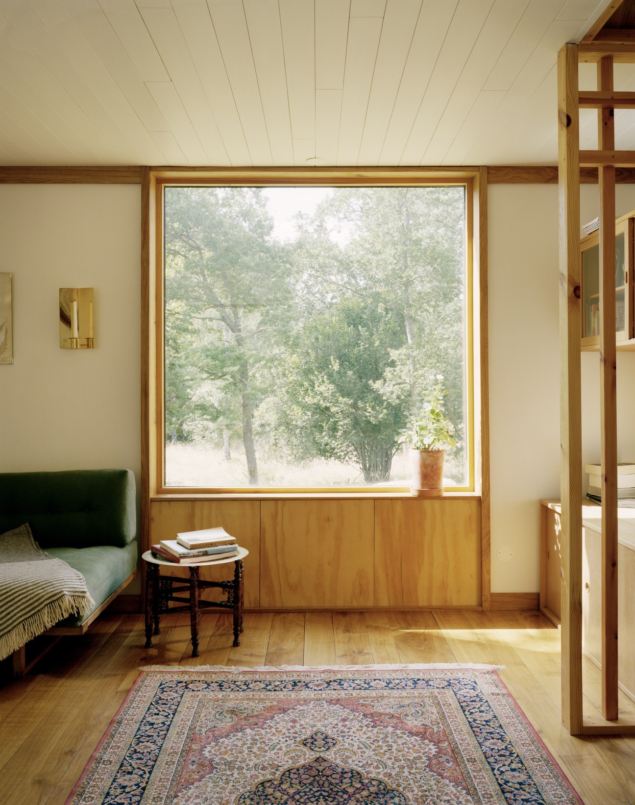 Bedroom with large square window framing forest views, wooden bed with green headboard, and patterned rug on oak floor.