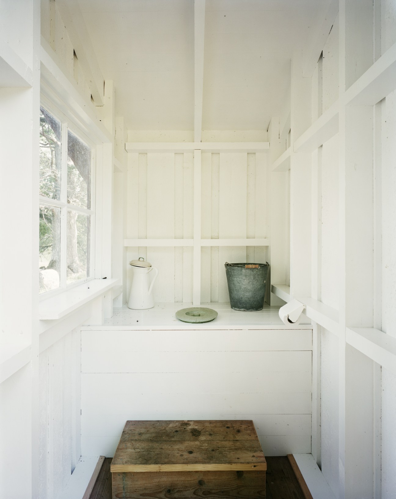 Minimalist white toilet hut interior with wooden seat, window, white pitcher, metal bucket and simple furnishings.