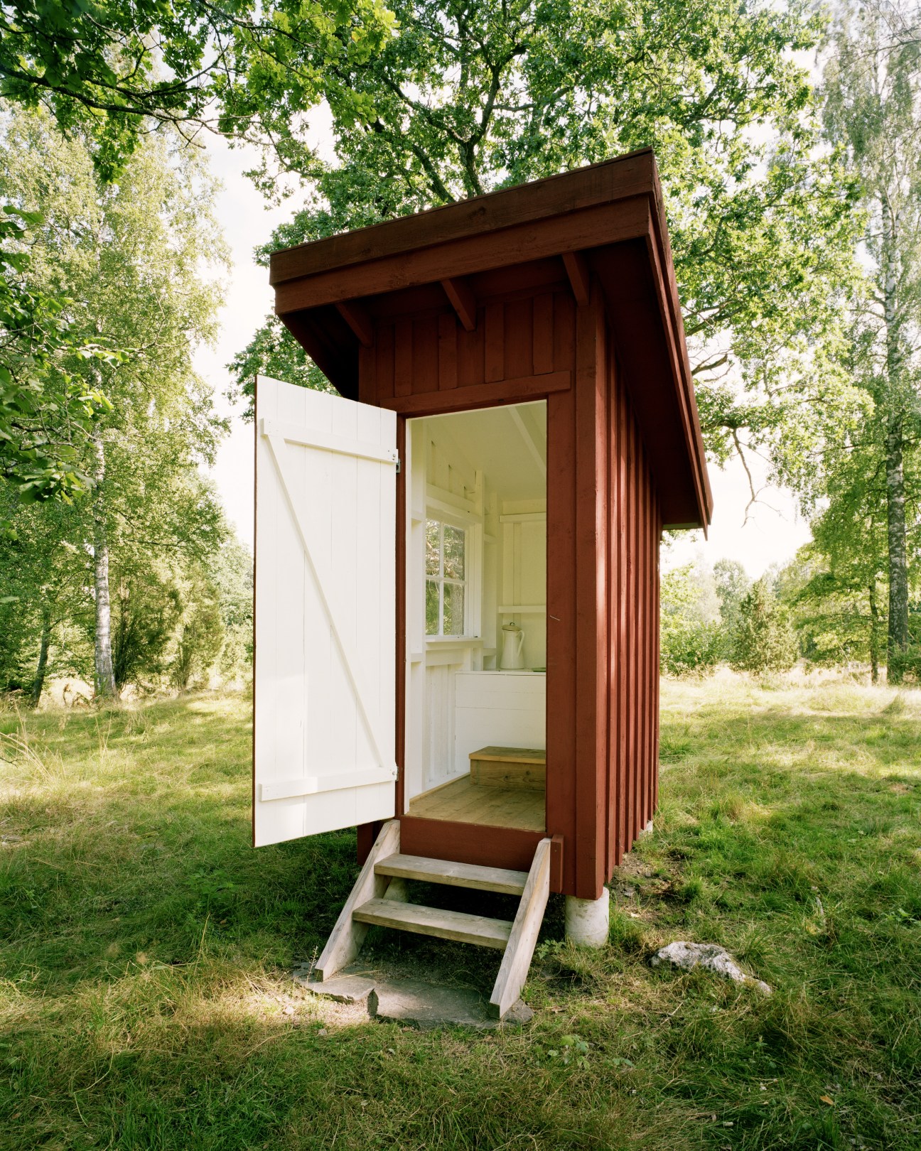 A dark reddish-brown wooden toilet hut with white interior visible through open door, surrounded by pine trees.