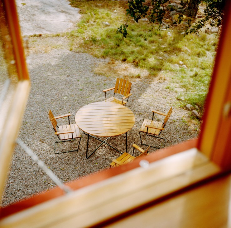 Grythyttan outdoor furniture arranged around a wooden table on gravel, viewed through a red window frame.