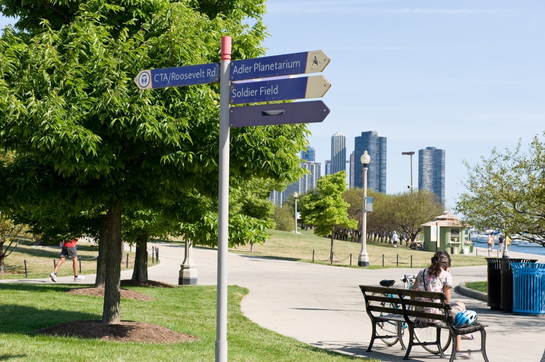 Chicago lakeside park path with directional signs, trees, and skyline in background.