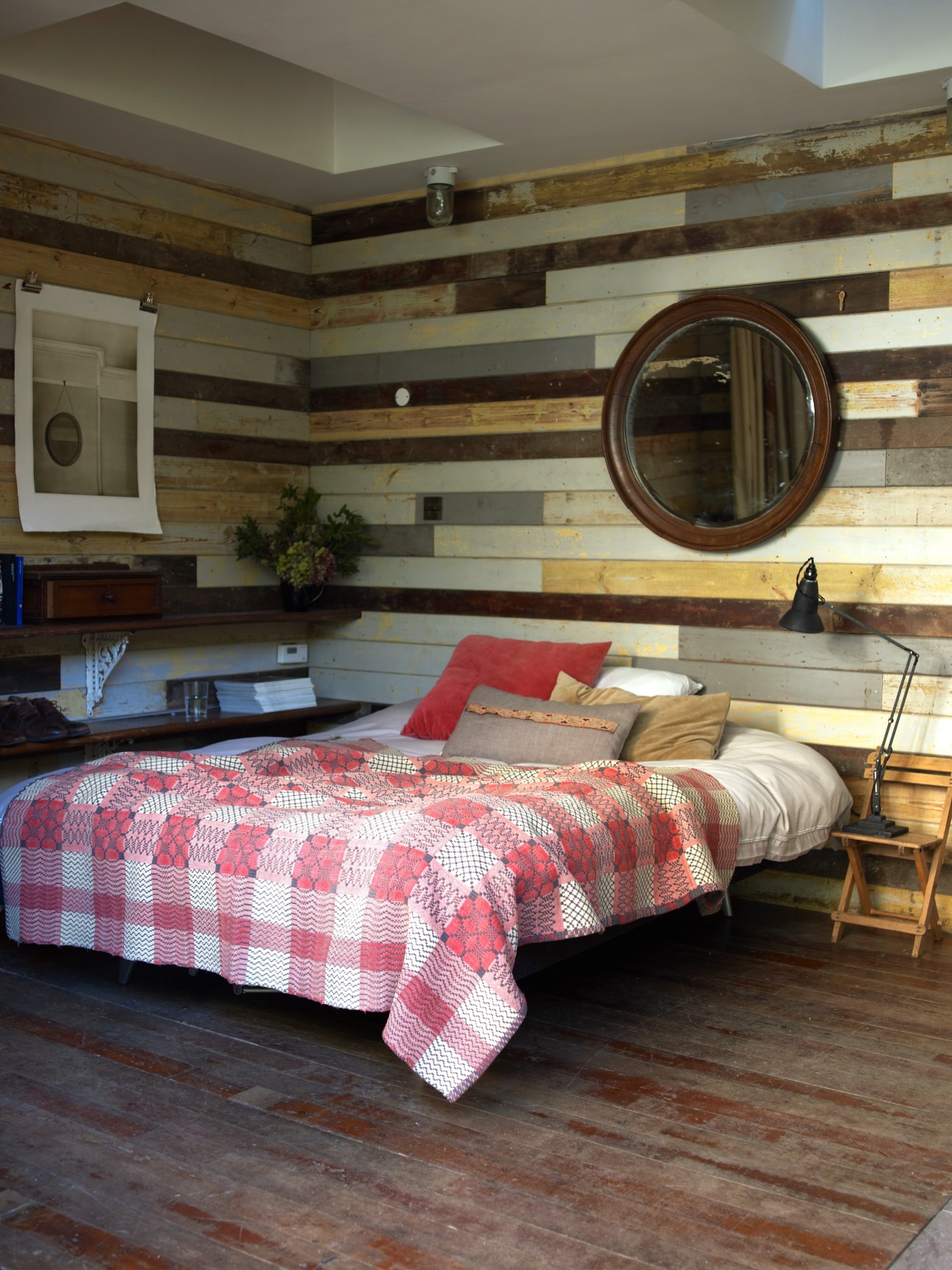 Bedroom with striped salvaged wood walls, red patterned quilt, and circular mirror in garden cabin by Retrouvius.