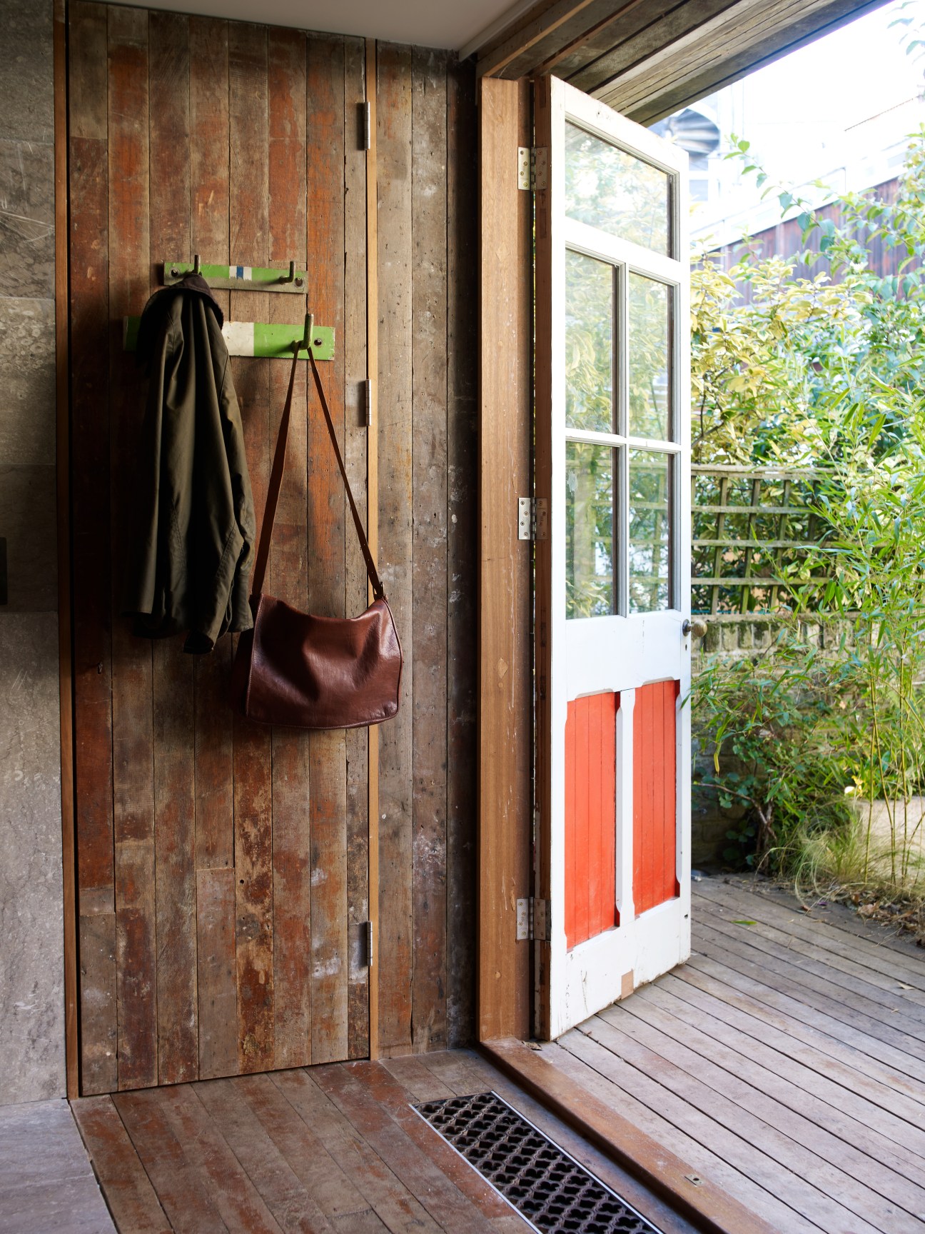 Interior of garden cabin with reclaimed wooden wall, green coat hooks, brown leather bag, and open door to garden.