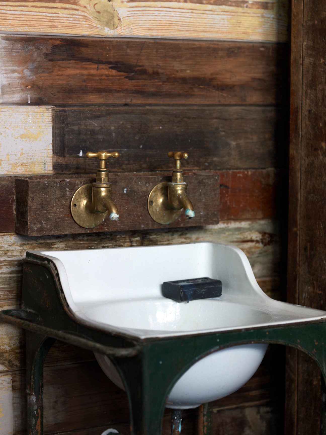 Vintage bathroom sink with white basin, brass taps, and weathered wooden wall paneling in the garden cabin designed by Retrouvius.