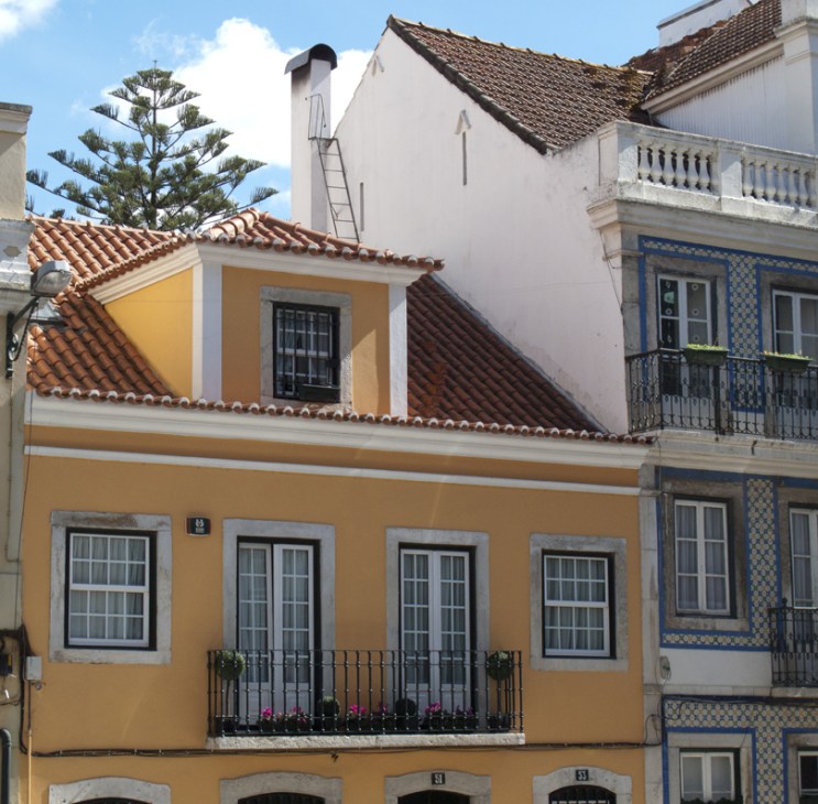 Yellow-painted Pombaline-style building with terracotta tile roof, blue-tiled neighbor, and palm tree in Lapa.