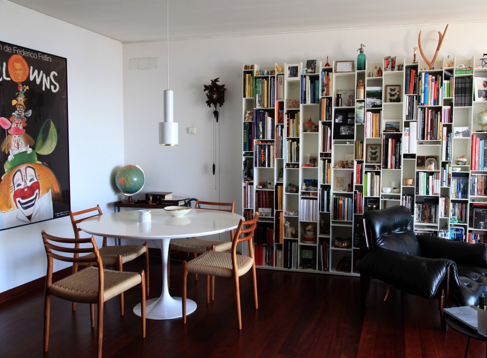 Modern living room with round white table, orange mid-century chairs, floor-to-ceiling bookshelves, and Fellini film poster.