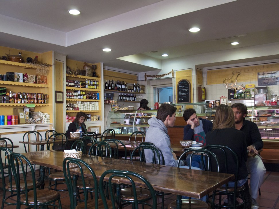 Interior of Chef restaurant in Lapa with customers seated at wooden tables and green bentwood chairs, yellow shelving with bottles and products behind counter.