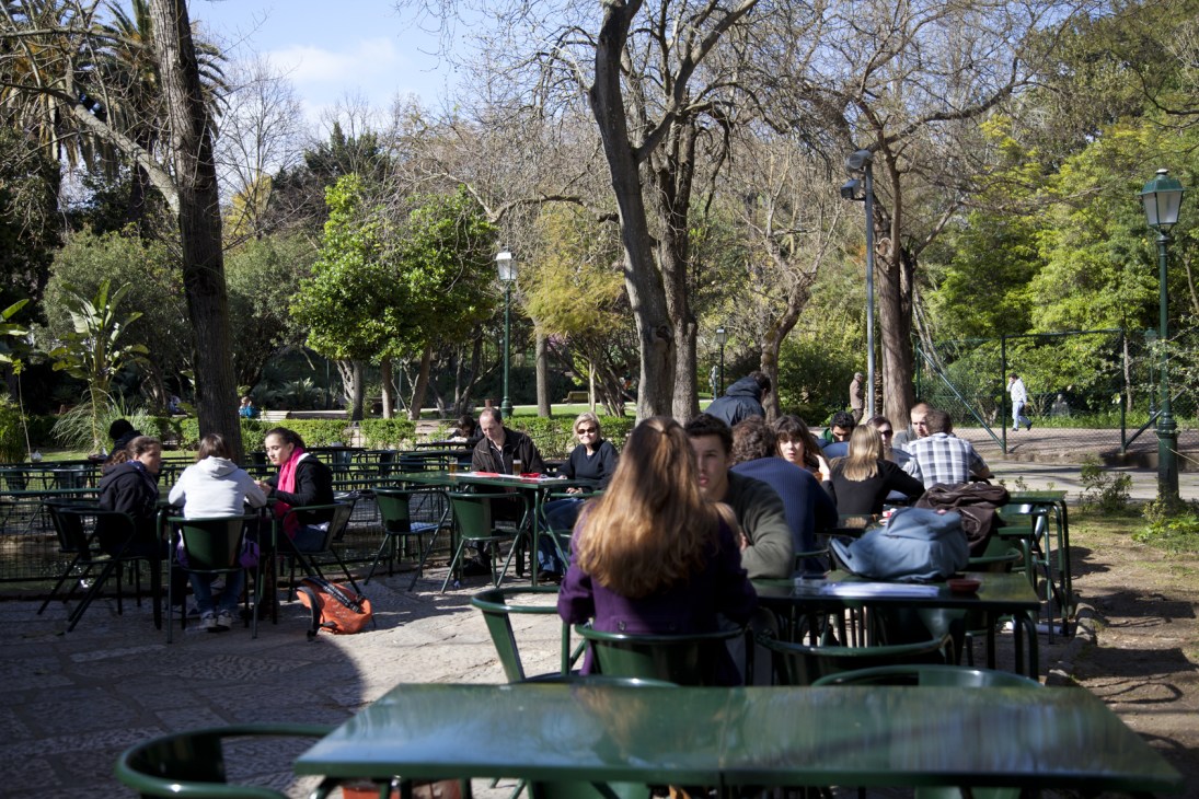 People sitting at green tables in a sunny park surrounded by tall trees.