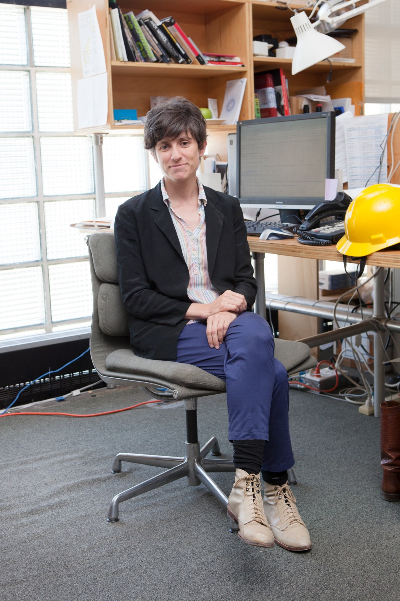 Curator Larissa Harris sits in an office chair in a bright workspace with wooden shelving and a yellow hard hat on the desk.