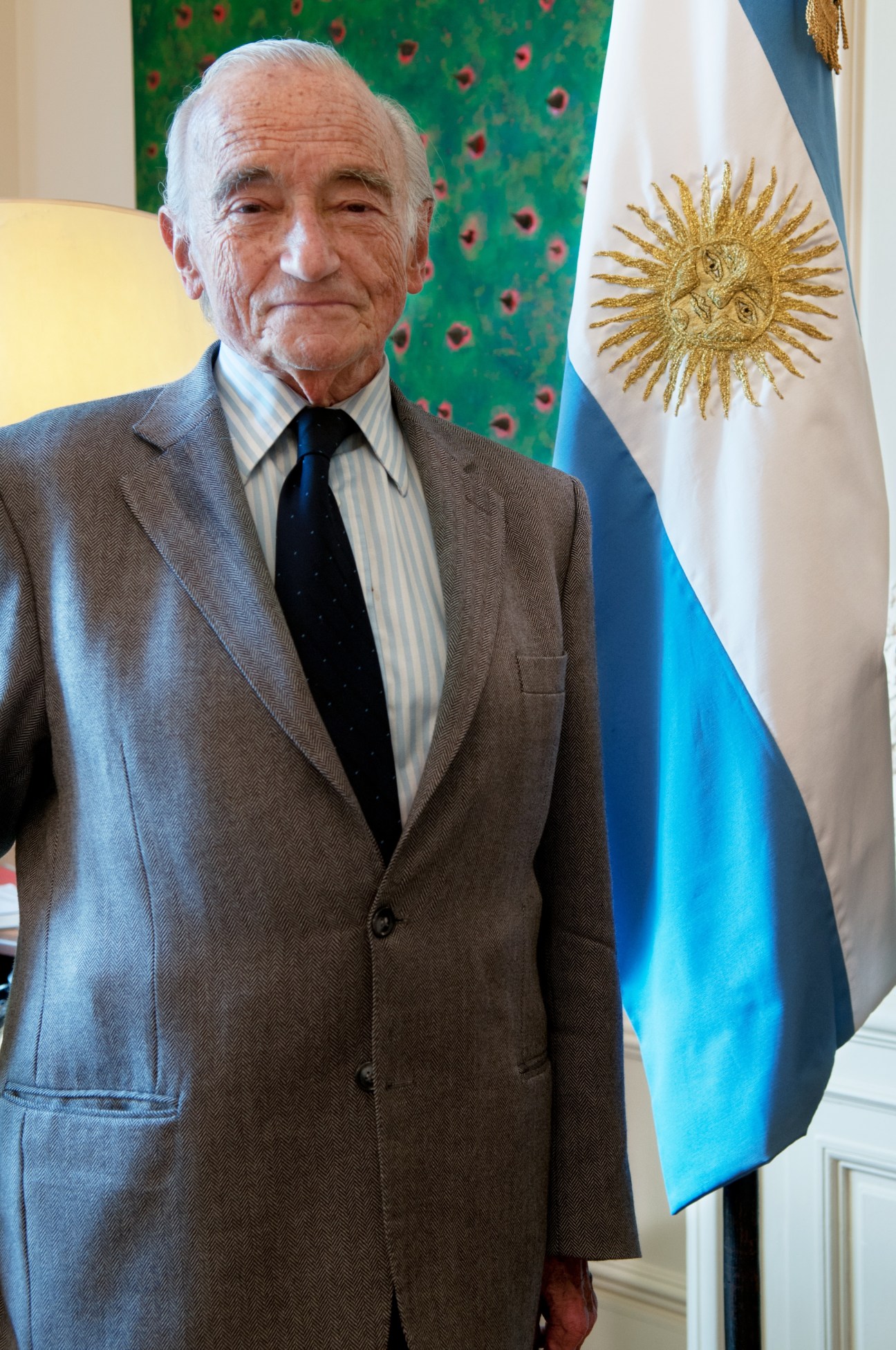 Elderly man in grey suit and dark tie standing beside Argentine flag with sun emblem.
