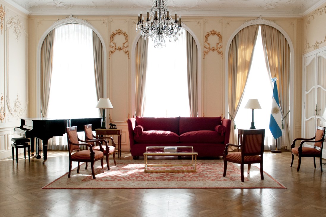 Elegant Parisian embassy reception room with burgundy sofa, piano, crystal chandelier and Argentine flag.