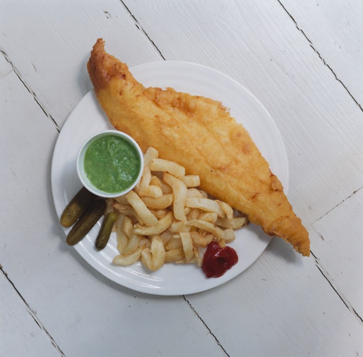 Fried haddock with chips, mushy peas, gherkins, tomato ketchup and vinegar on a white plate.