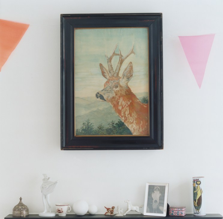 Framed painting of a deer with antlers hung on white wall above shelf with decorative objects.