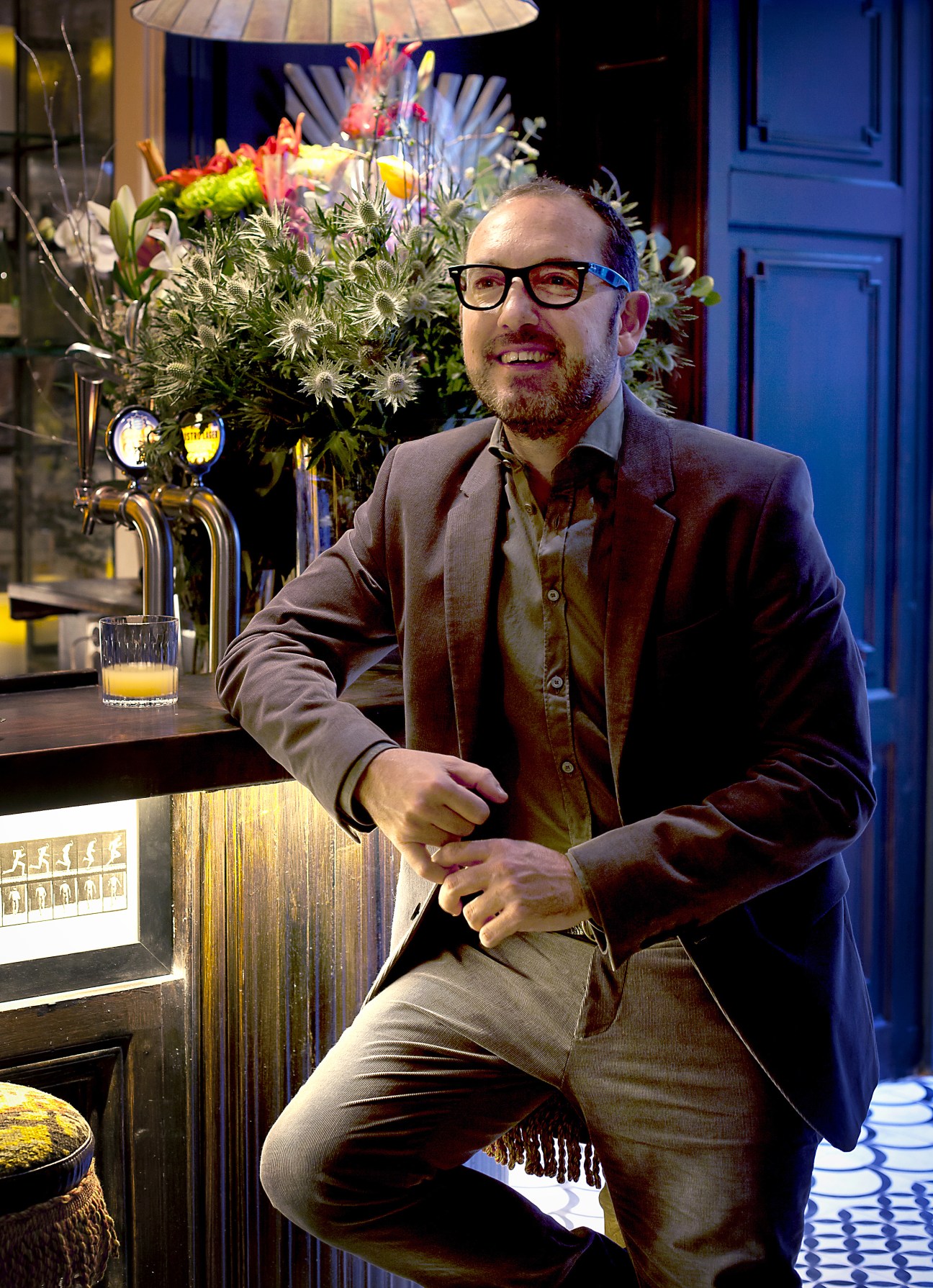 Catalan architect Lázaro Rosa-Violán sitting at a bar with colorful flowers and drinks visible behind him.