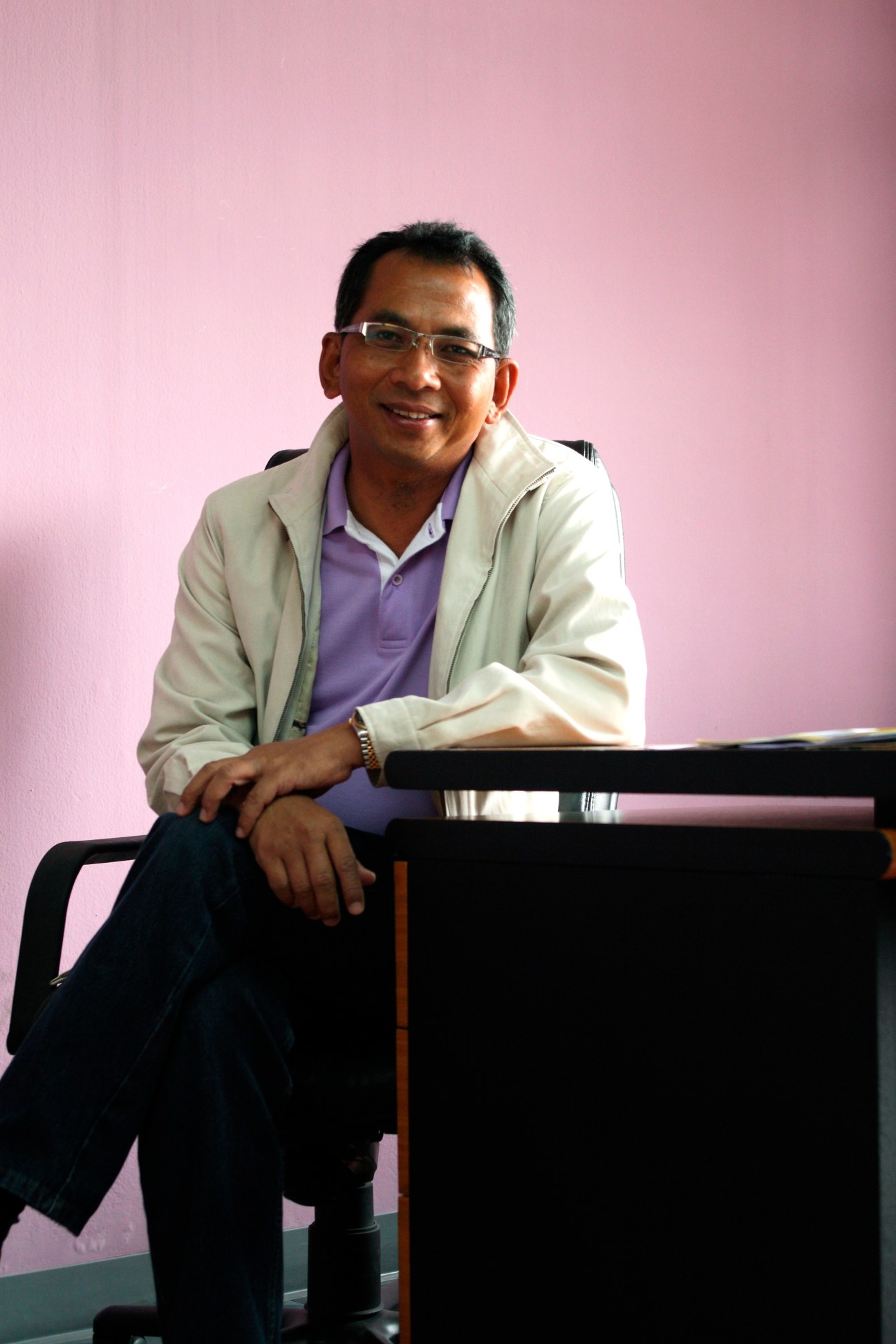 Sontiyan Chuenreuthainaidhama, founder of RDCK radio station, smiles while leaning against a desk against a pink background.