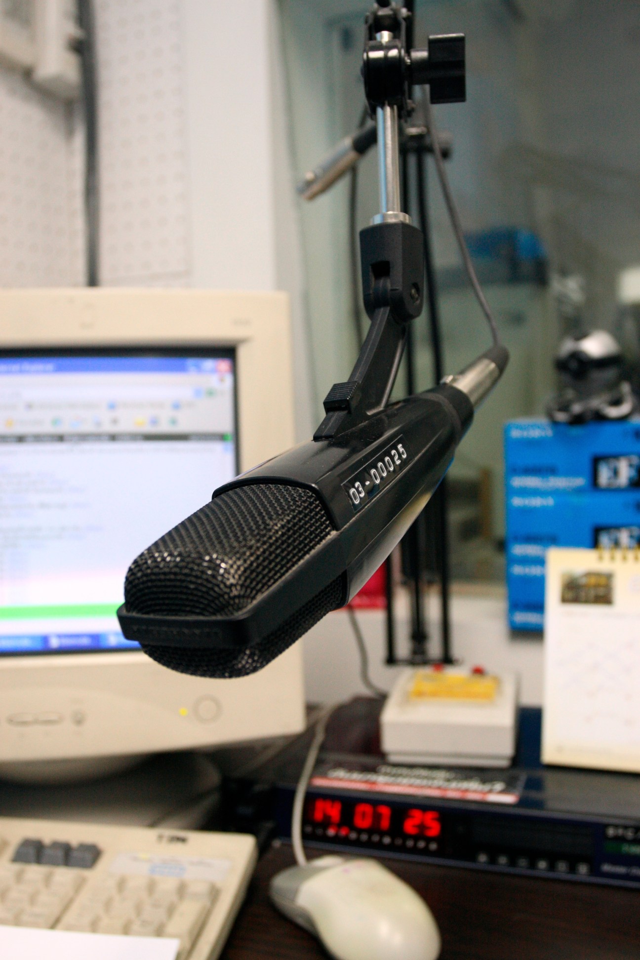 A professional microphone mounted on an articulated arm in a radio station studio with computer equipment visible in the background.