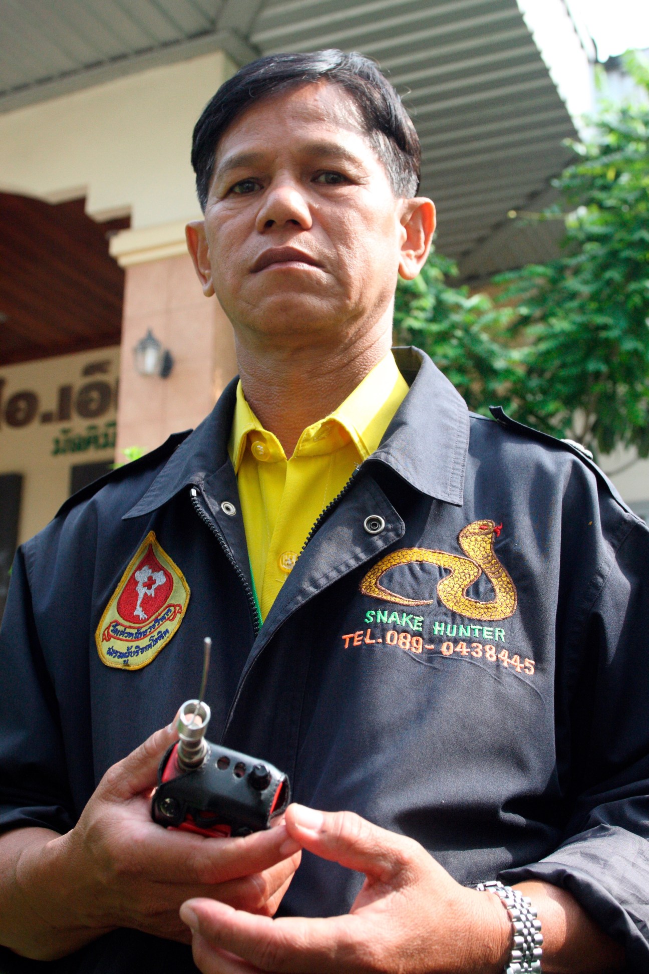 Sompop Sridarachop, a 54-year-old snake wrangler, stands outdoors wearing a black jacket with snake embroidery, holding a radio device.