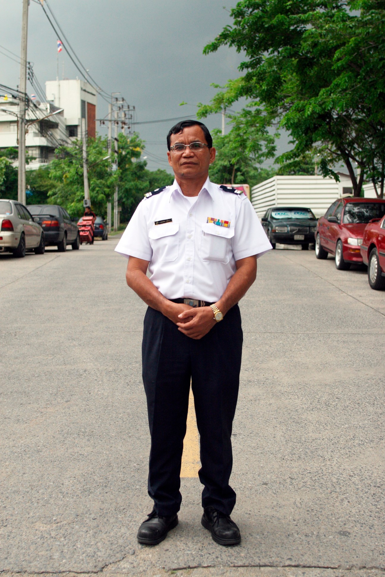 Prasat Sutthowa, a bus driver in white uniform shirt and dark trousers, stands on a Bangkok street lined with parked cars and buildings.