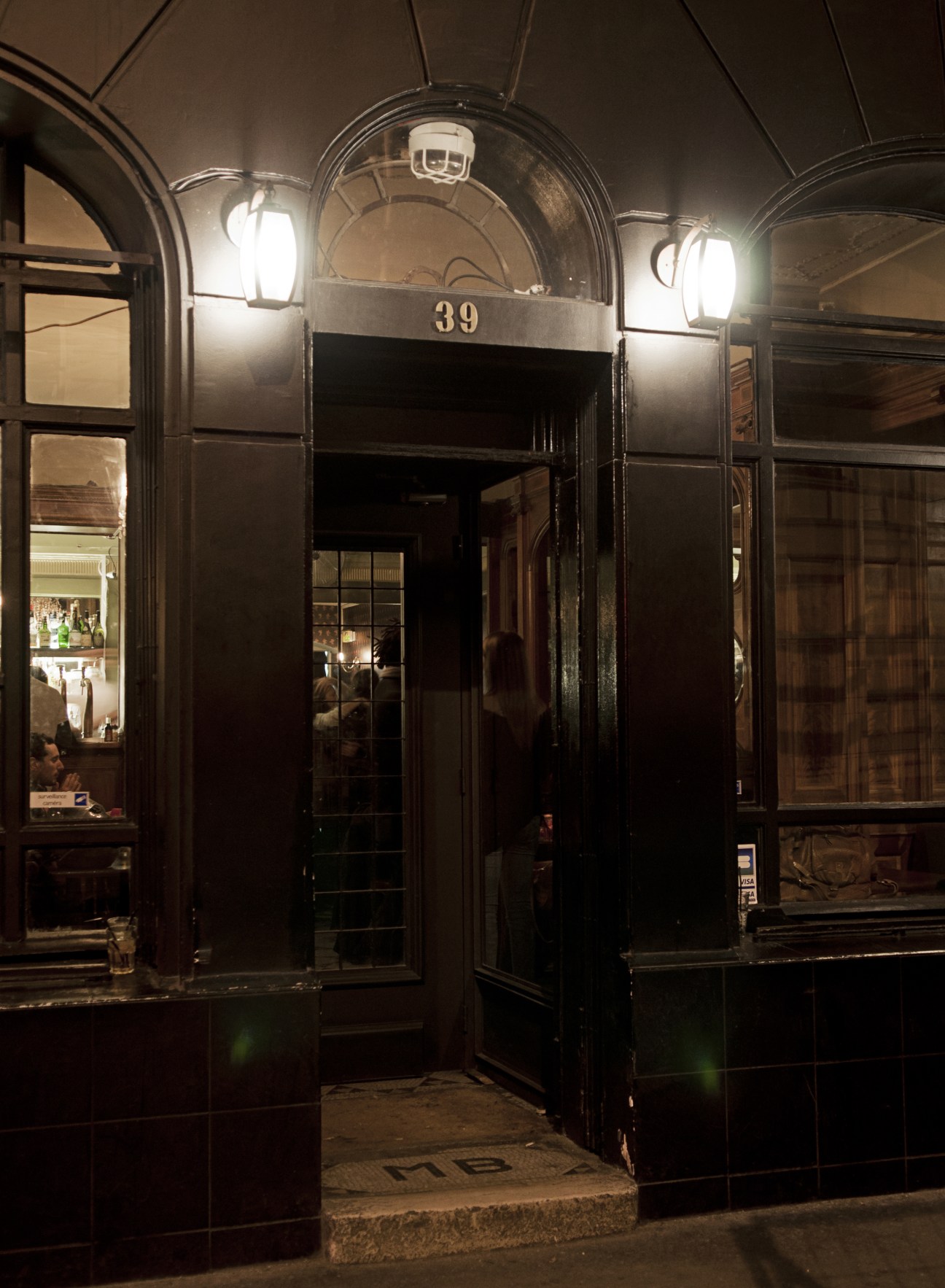 Nighttime exterior of Le Pompon bar entrance with dark wood facade, arched doorway, number 39, and illuminated sconces.
