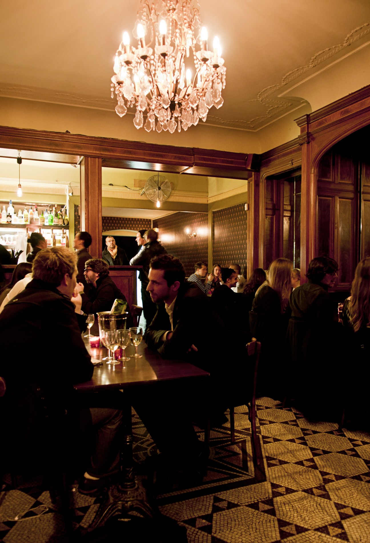 Patrons enjoying drinks in the elegant interior of Le Pompon bar with crystal chandelier and wood paneling.