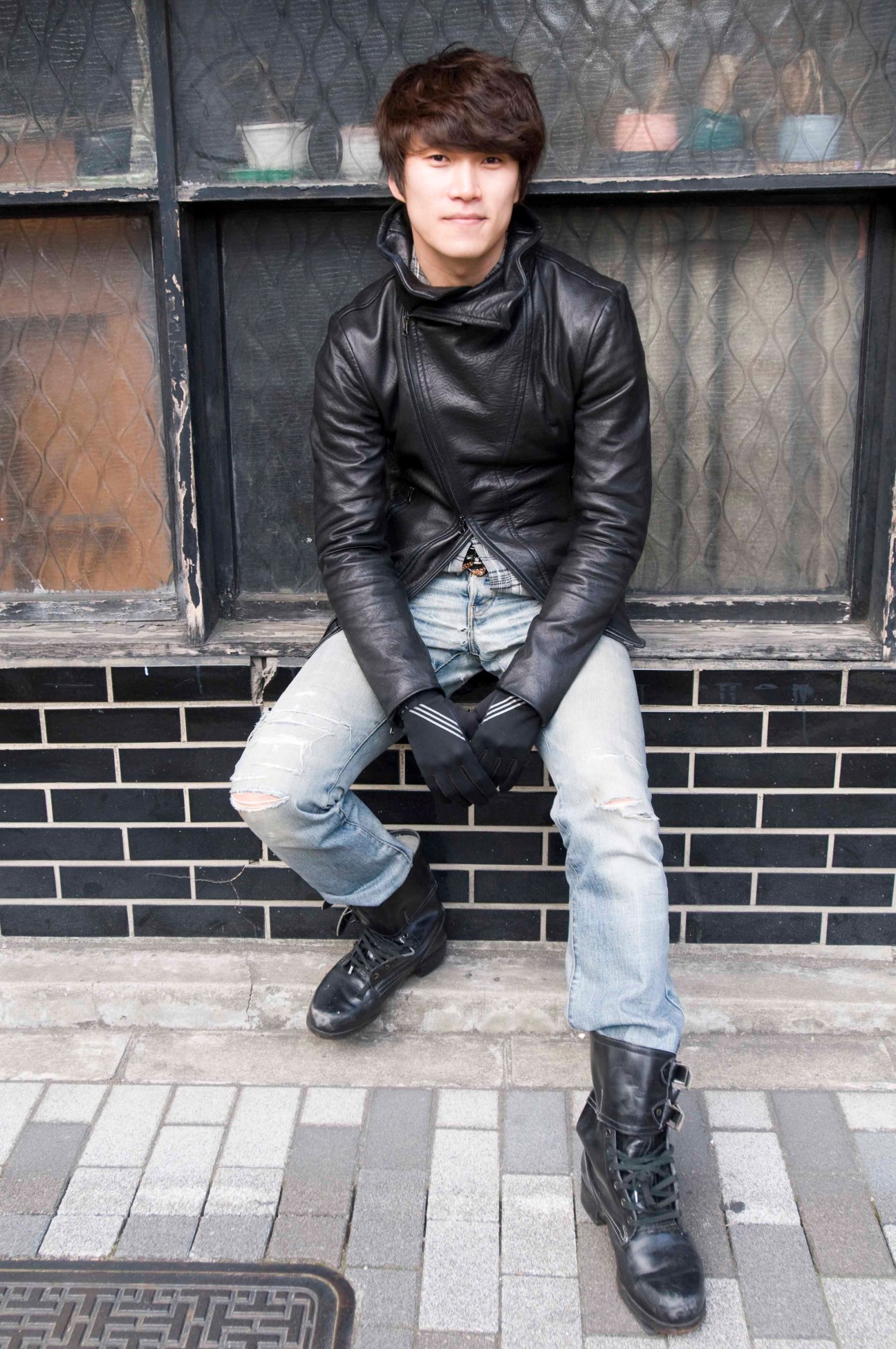 Choreographer Park Nam Yong sitting against a brick wall wearing a black leather jacket and ripped jeans.