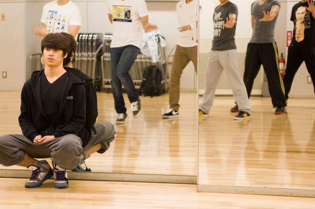 Choreographer Park Nam Yong crouches in front of a mirror while 2PM band members rehearse dance moves behind him in a studio.