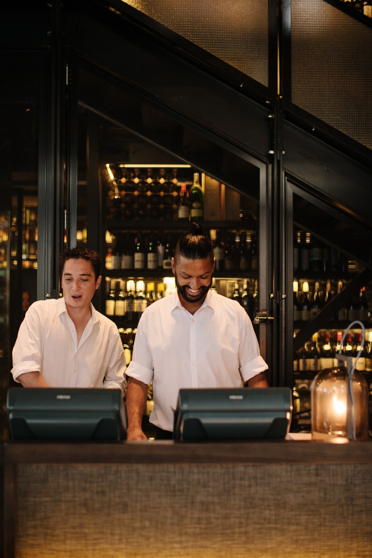 Two bartenders in white shirts working at a bar with illuminated bottle display behind them.