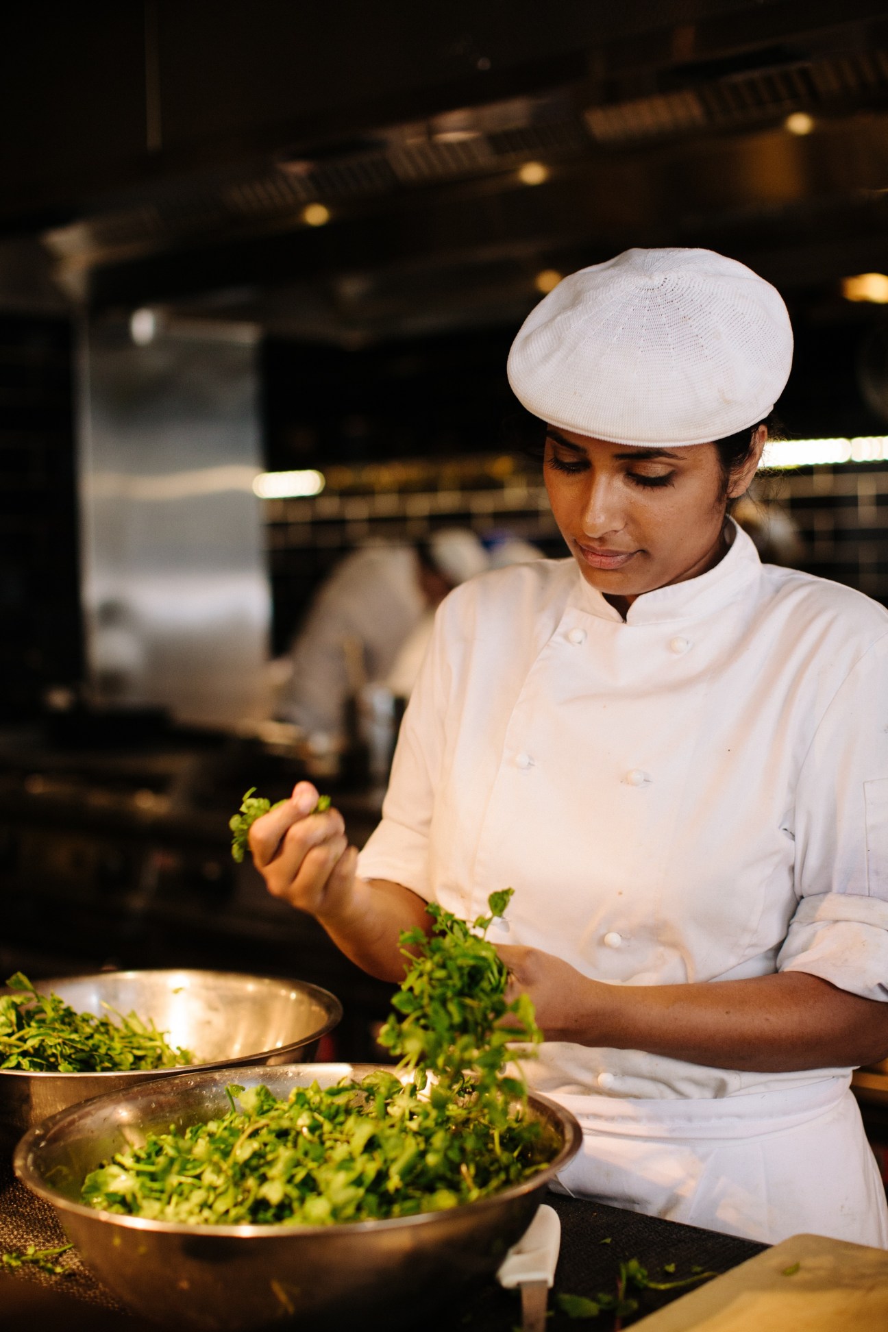 Chef in white chef's coat and hat preparing greens in a pan in an open kitchen.