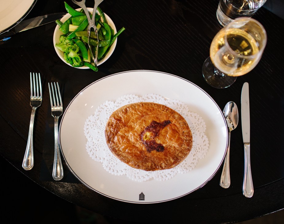 Seafood pie with puff pastry crust served on white plate with green vegetables and wine glass.