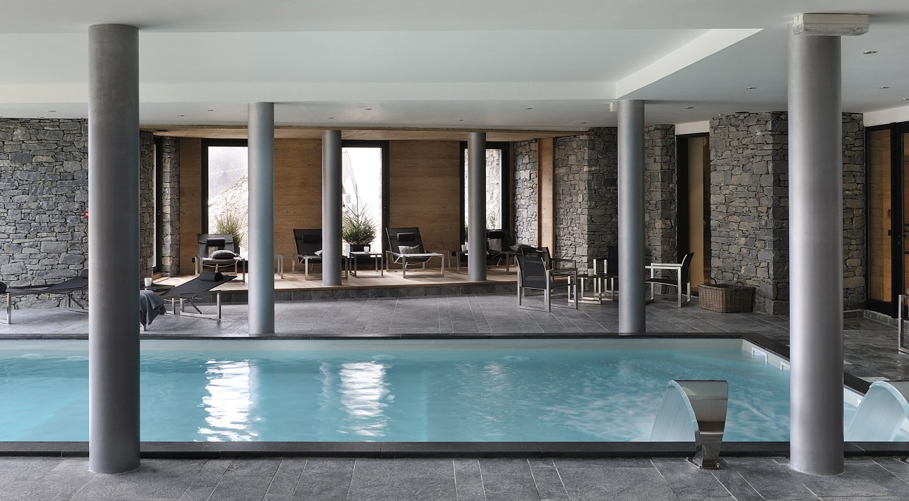 Indoor pool with stone walls, concrete columns, and seating area at Les Granges d'en Haut hotel in Chamonix.