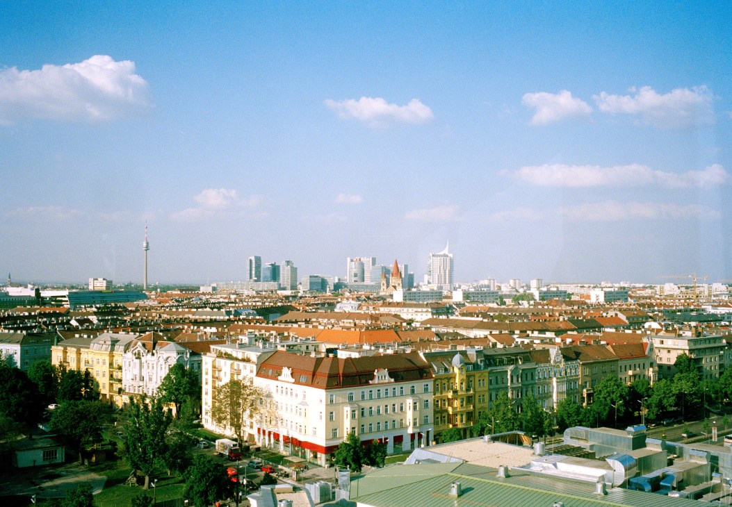 Vienna's cityscape with traditional low-rise buildings in foreground and modern high-rises and TV tower visible in the distance under clear blue sky.