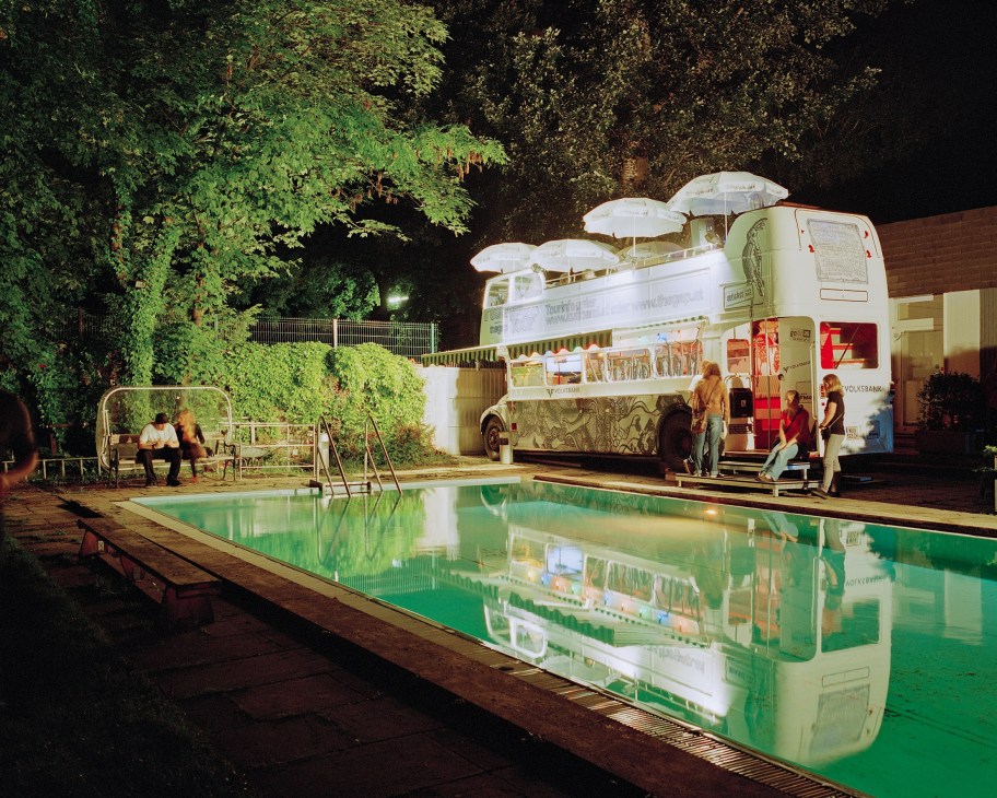 A vintage double-decker bus converted into a bar overlooks an illuminated green pool at night, surrounded by trees and lounging visitors.