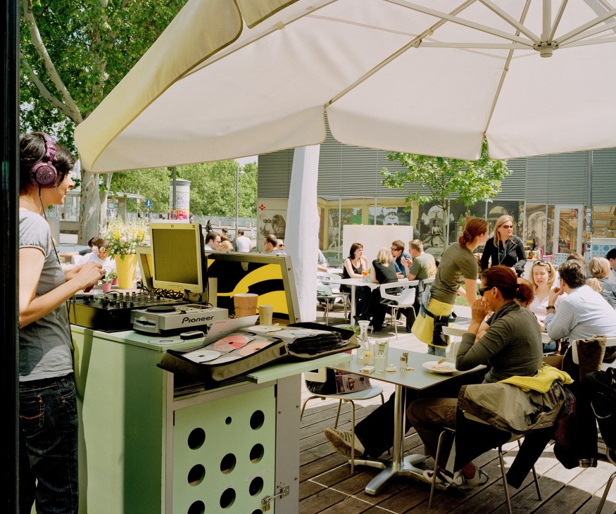 DJ performing under a white umbrella at an outdoor café with diners seated at tables.