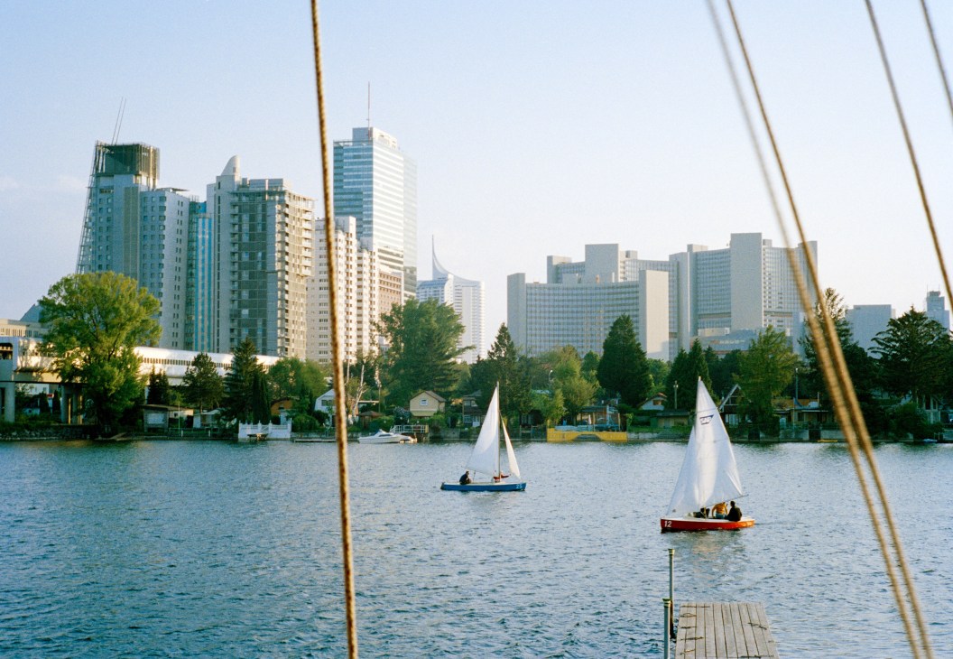 Small sailboats on the Danube river with Vienna's modern skyline and trees visible across the water.