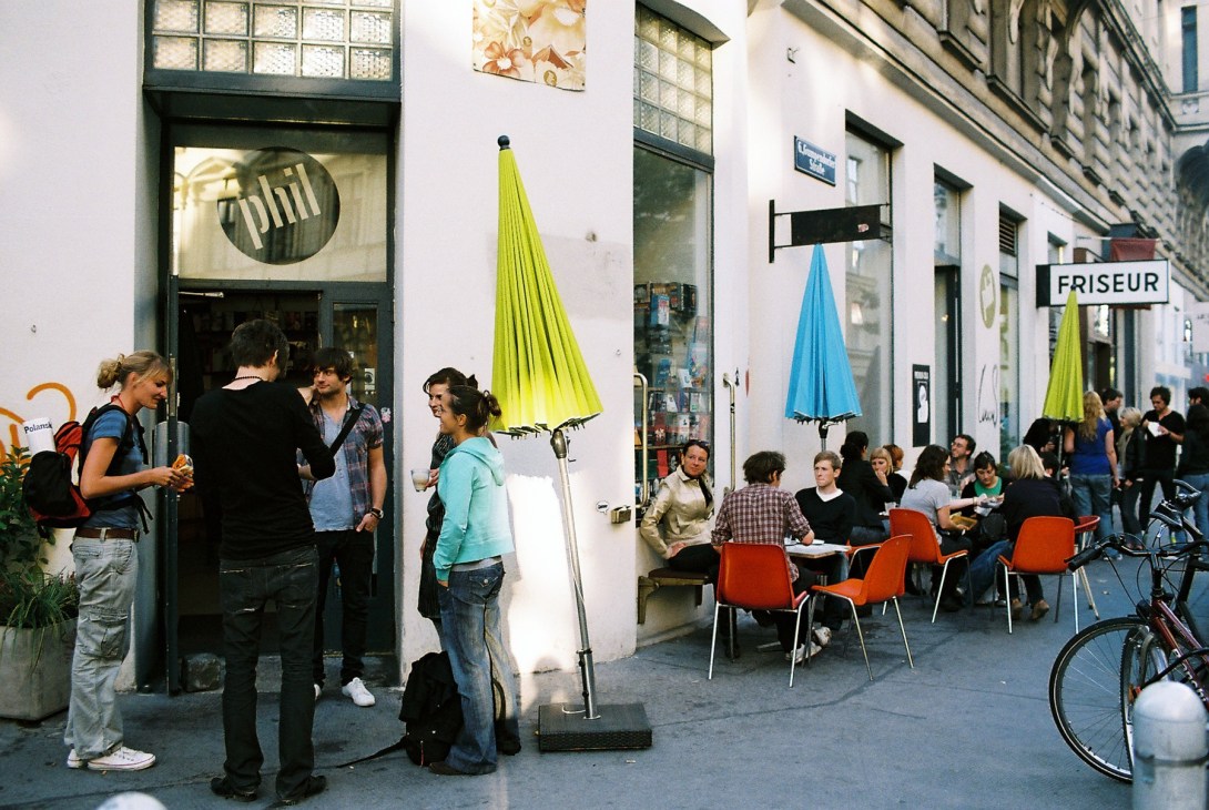 Street scene outside Phil bookshop with people gathering, colorful umbrellas, and outdoor seating on a Vienna street.