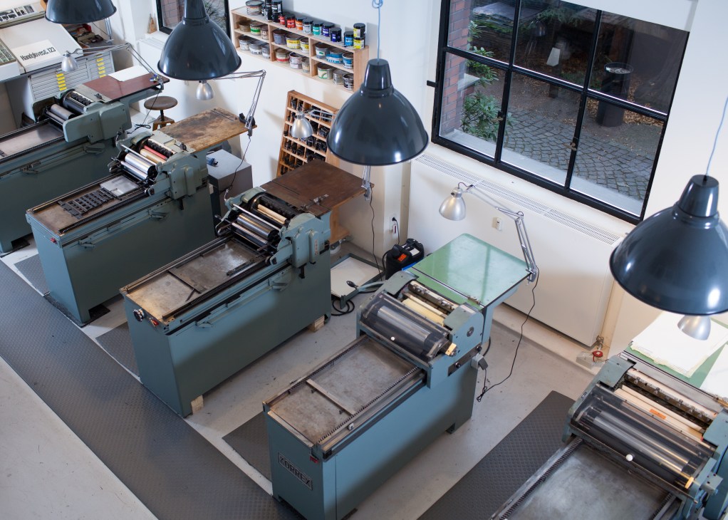 Overhead view of Korrex letterpress machines in a bright workshop with shelving and black pendant lamps.
