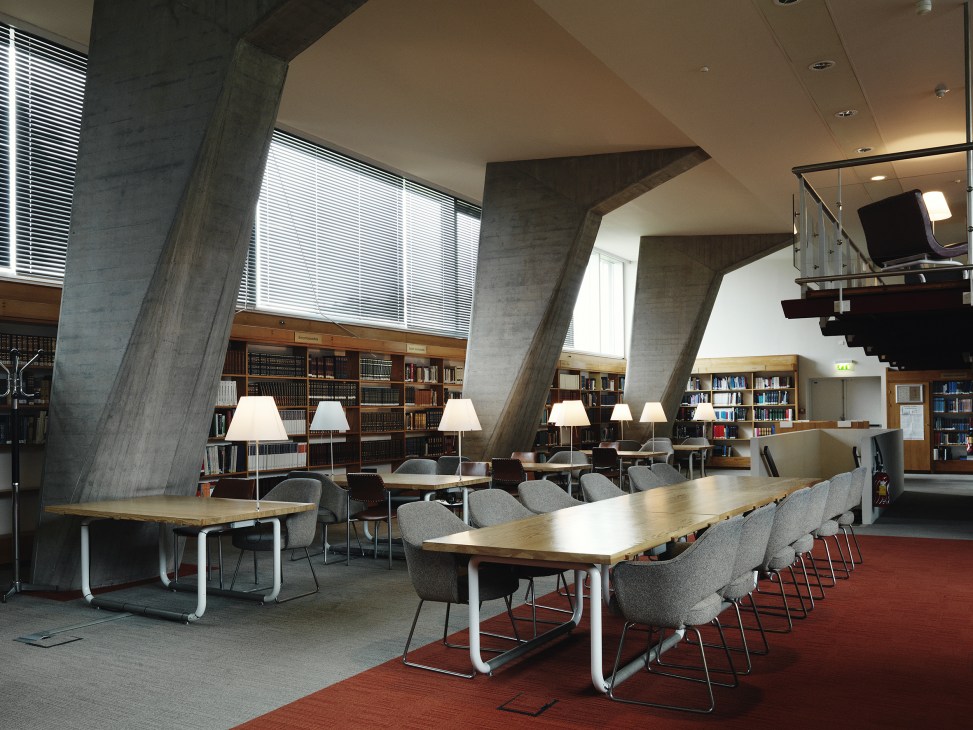 Modernist library interior with concrete columns, wooden bookshelves, reading tables and grey chairs.