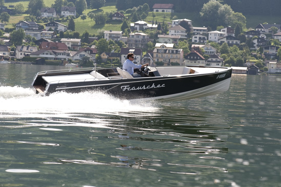 Man operates Frauscher Lido 686 motor boat on Alpine lake with lakeside village in background.