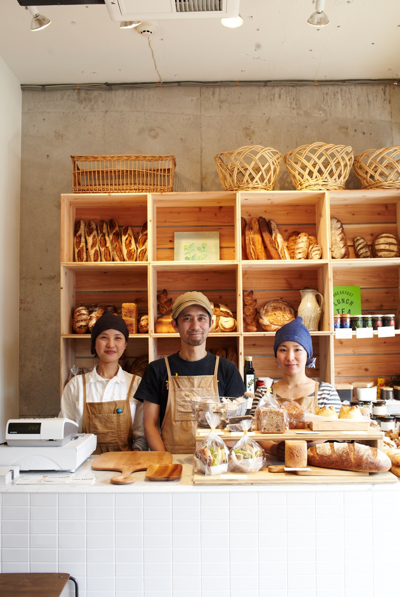 Three staff members in aprons stand behind a bakery counter displaying fresh bread and pastries.