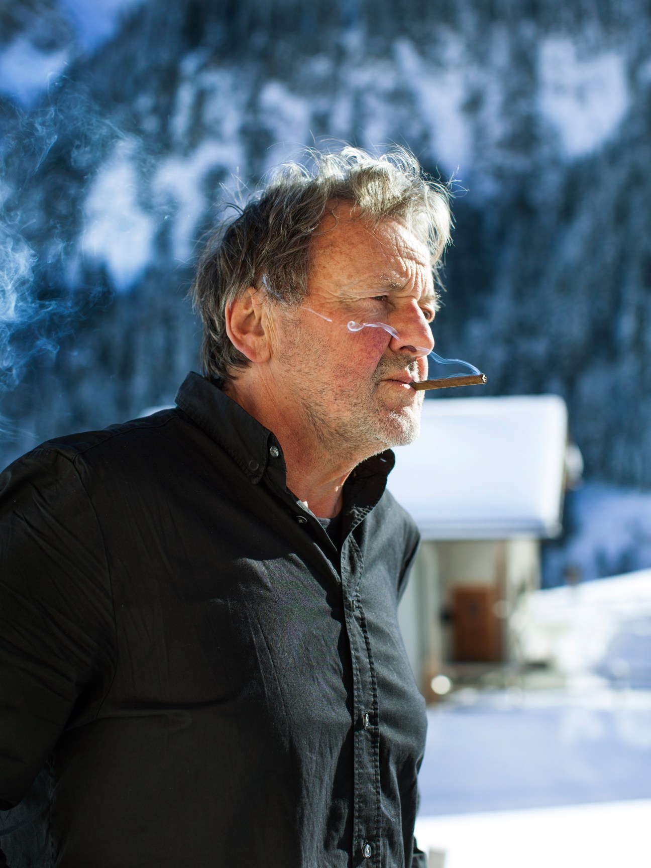 Swiss architect Gion Caminada smoking a cigar in profile with snowy Alpine mountains in the background.