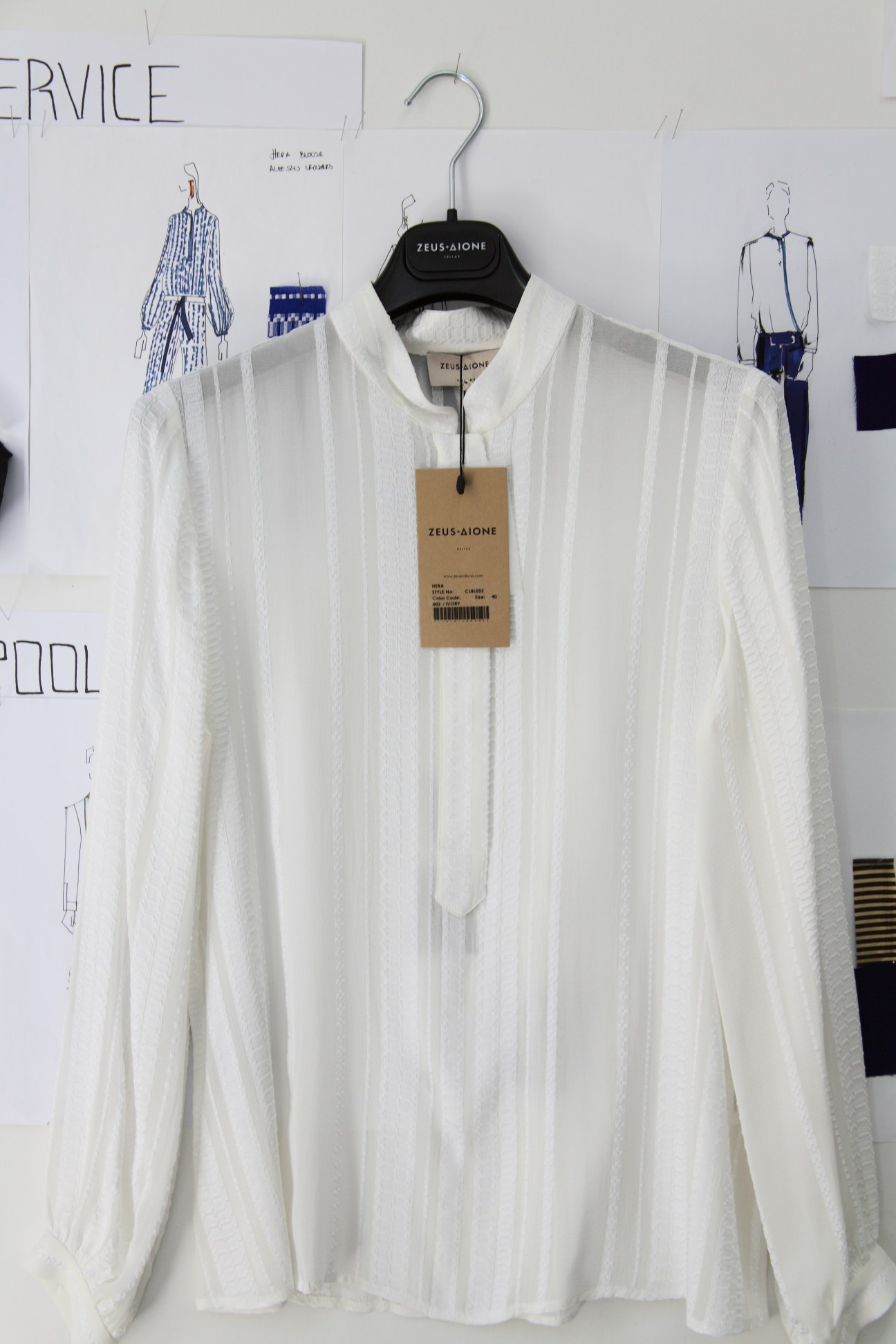 White linen tunic on hanger with Zeus + Dione label, displayed with uniform sketches on wall behind.