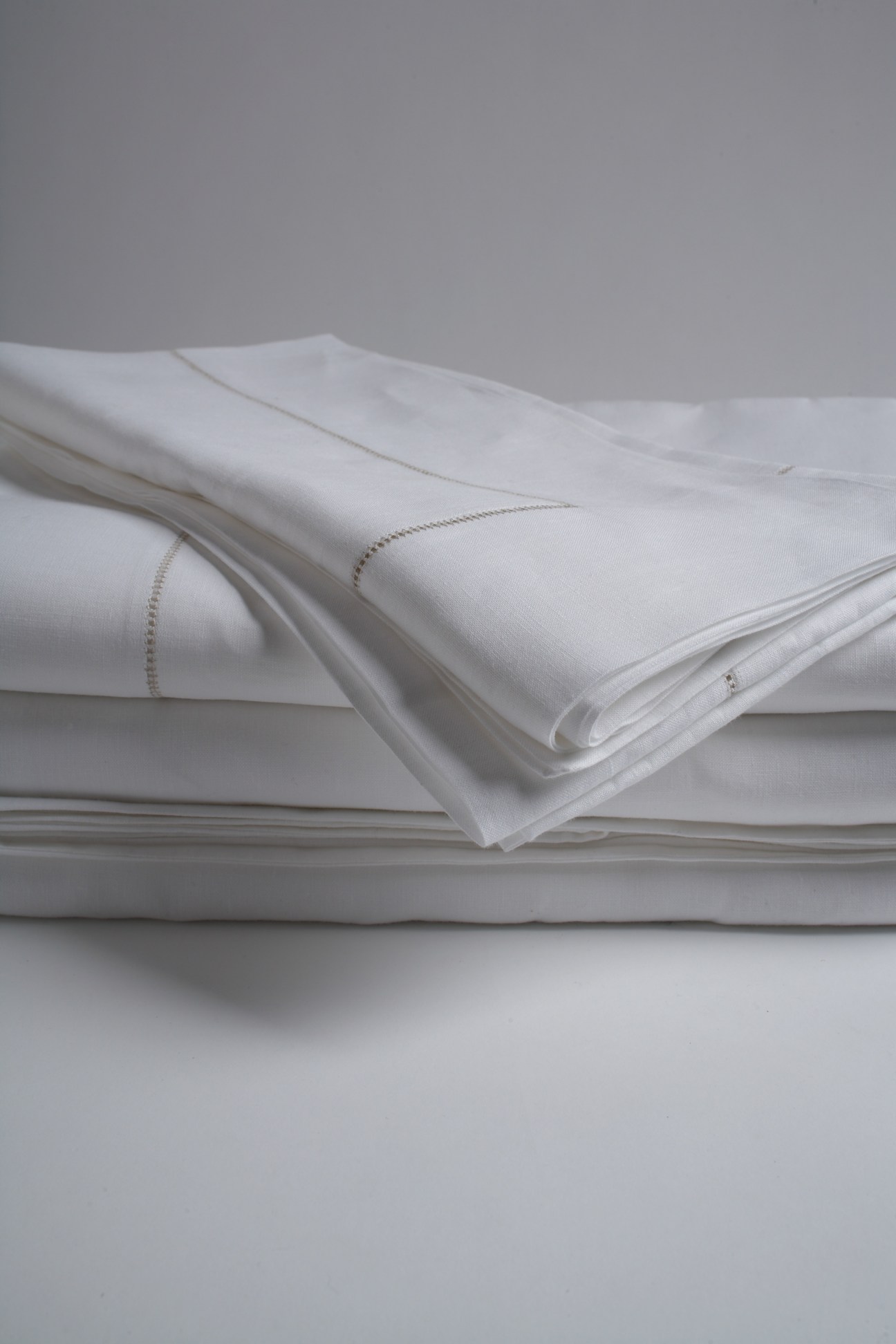 Folded white linen sheets with detailed stitching displayed on a neutral surface.