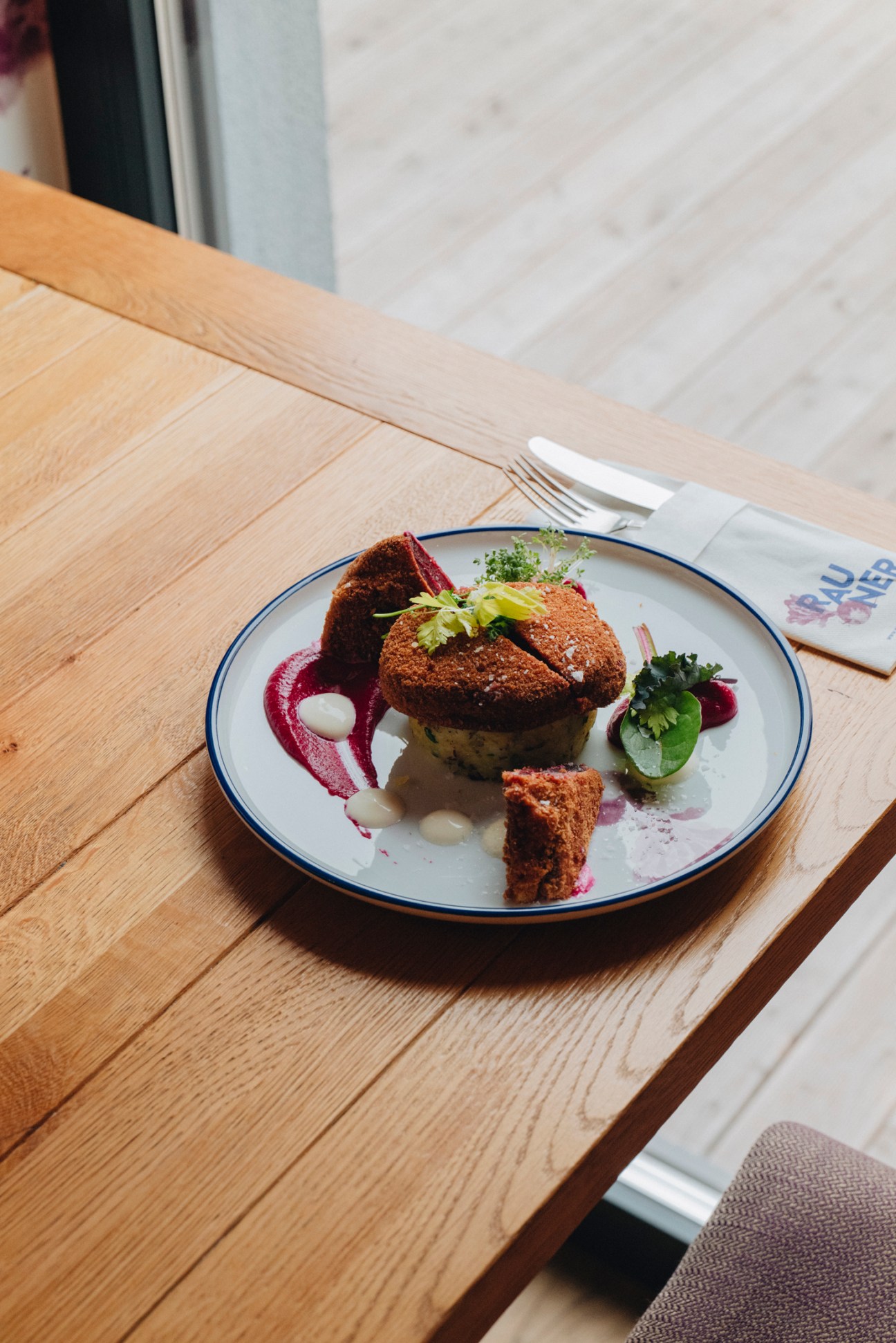 Elegantly plated beetroot dish with crispy coating and purple sauce on white plate with blue rim at wooden table
