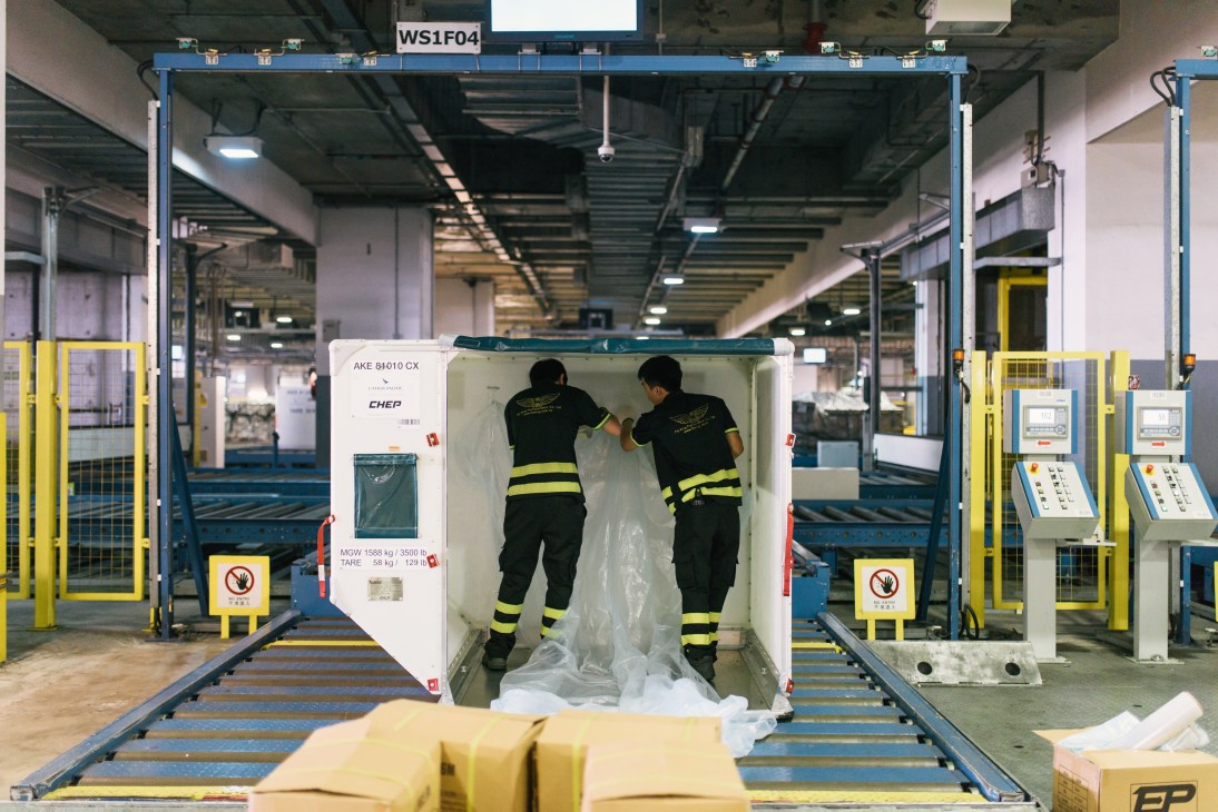 Two workers in safety gear load cargo into a white shipping container at Hong Kong International Airport's Quick Transhipment handling area.