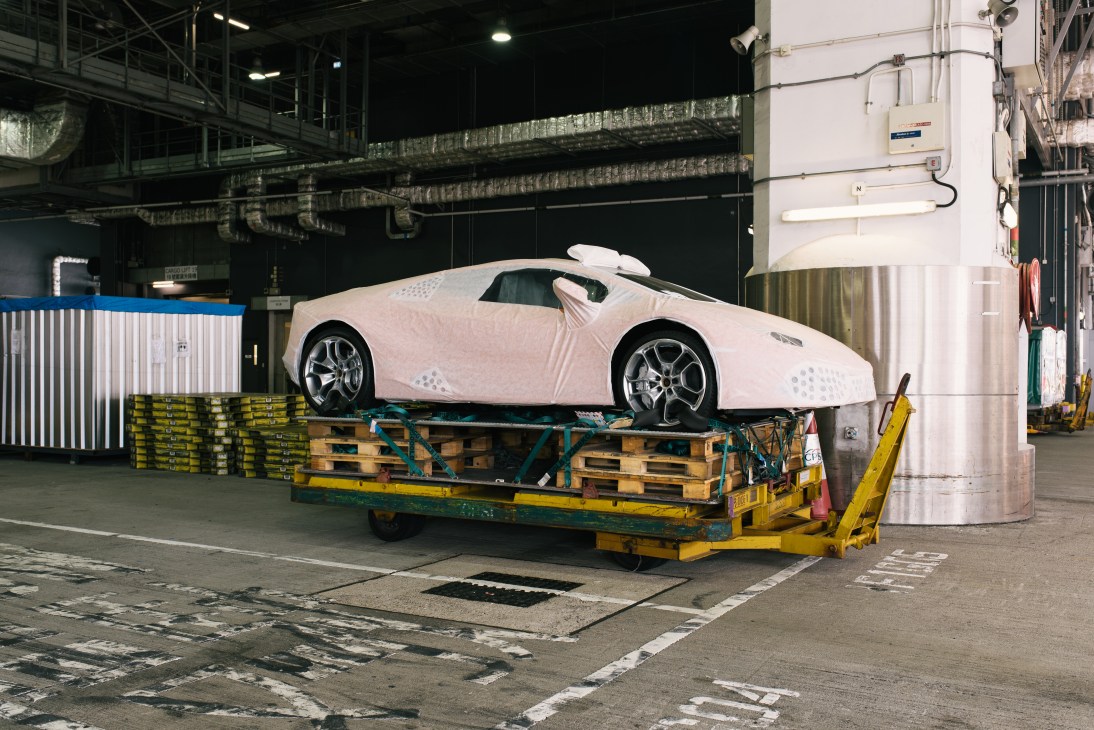 White Lamborghini sports car wrapped in protective plastic on yellow cargo pallet at Hong Kong International Airport.